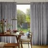 Full-length gray geometric pleated curtains, hung from a rod, pulled aside at center to reveal a sunlit backyard; wooden dining table with pancakes, chairs and pendant lamp occupy the foreground.
