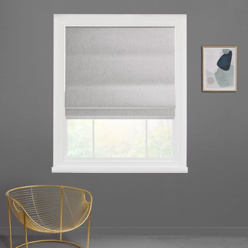 Gray fabric Roman shade lowered partway, smooth folds filtering diffuse daylight through window; white-trimmed opening in a gray wall with gold wire chair and abstract wall art.