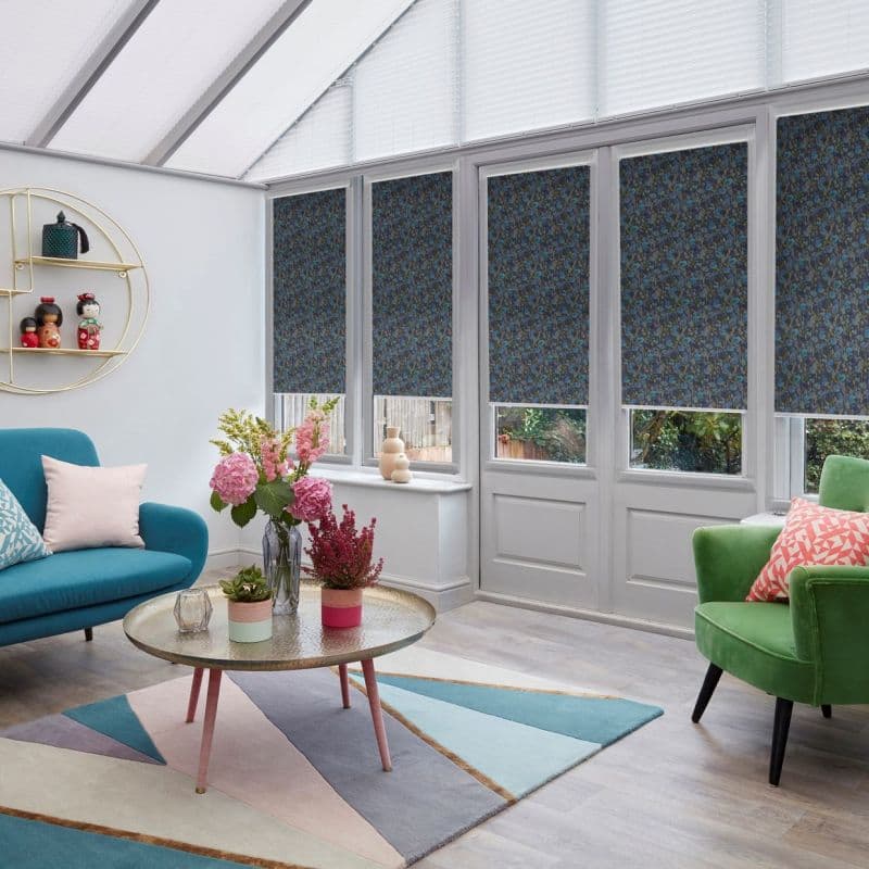 Blue floral roller blinds lowered across tall conservatory windows and doors, covering most panes; partially closed, filtering daylight into a bright sunroom with pitched glass roof, furniture and garden view.