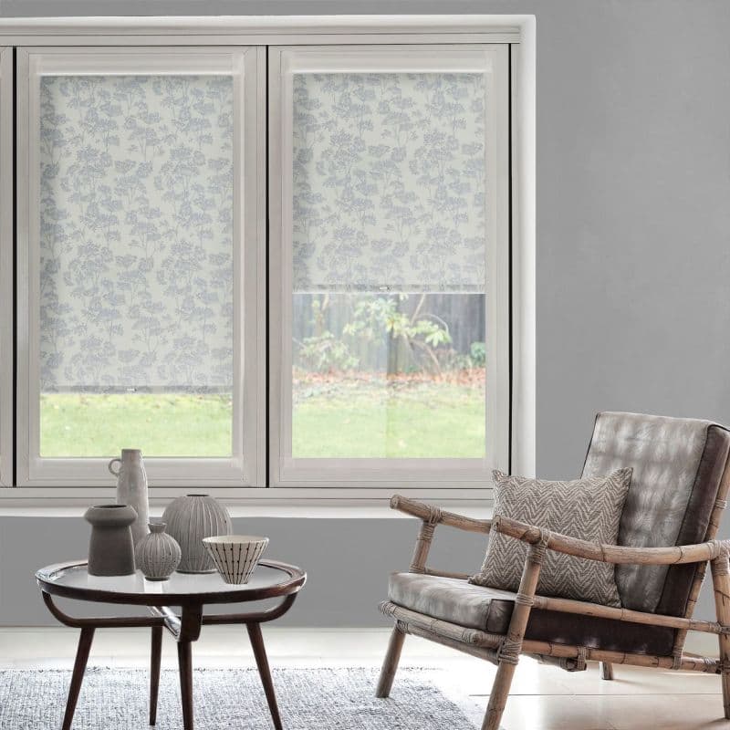 Floral-patterned roller blinds on a double window, one fully lowered and one partially raised, filtering soft daylight; living room with grey walls, rattan armchair, round coffee table, and garden view.