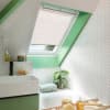 Pleated skylight blind in pale pink, mounted inside a roof window and lowered to filter daylight; sloped-ceiling bathroom with mint-green trim, white herringbone tiles, sink, bathtub and potted plant.