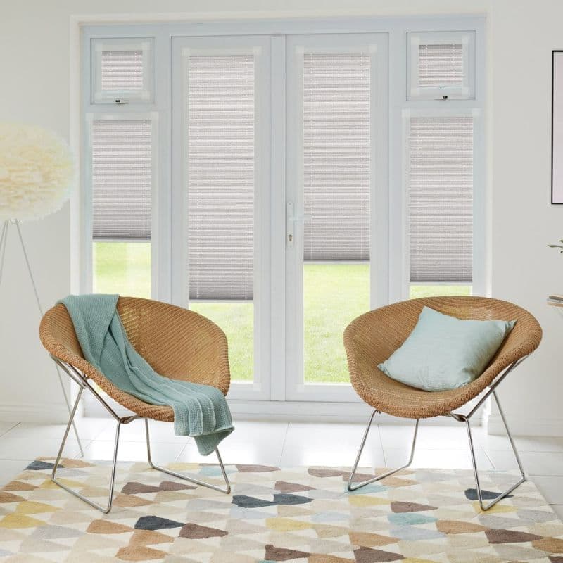 Pleated gray cellular blinds covering French doors and side windows, partially lowered and filtering daylight. Two wicker lounge chairs with mint textiles face the doors in a bright sitting area.