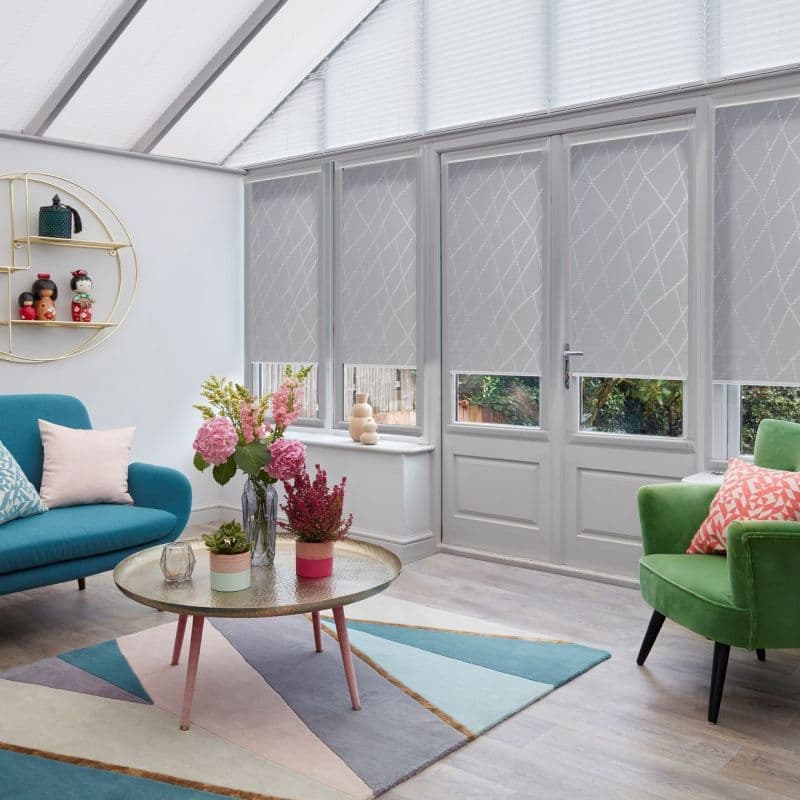Patterned grey roller blinds covering tall conservatory windows and door, lowered to mid-height and diffusing light; living room with teal sofa, green armchair, rug, coffee table, floral arrangements.