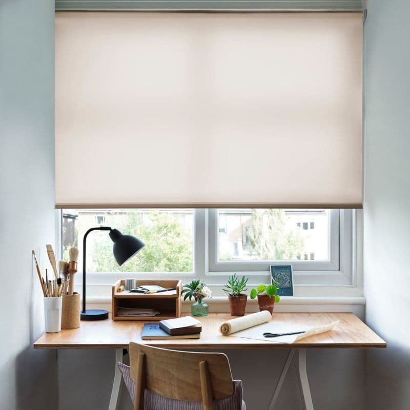 Object: Roller blind — beige, smooth fabric lowered over the window. Action: filtering soft daylight through a narrow gap at the sill. Context: tidy wooden desk workspace with lamp, brushes, potted plants, outside trees and buildings. British Herbs