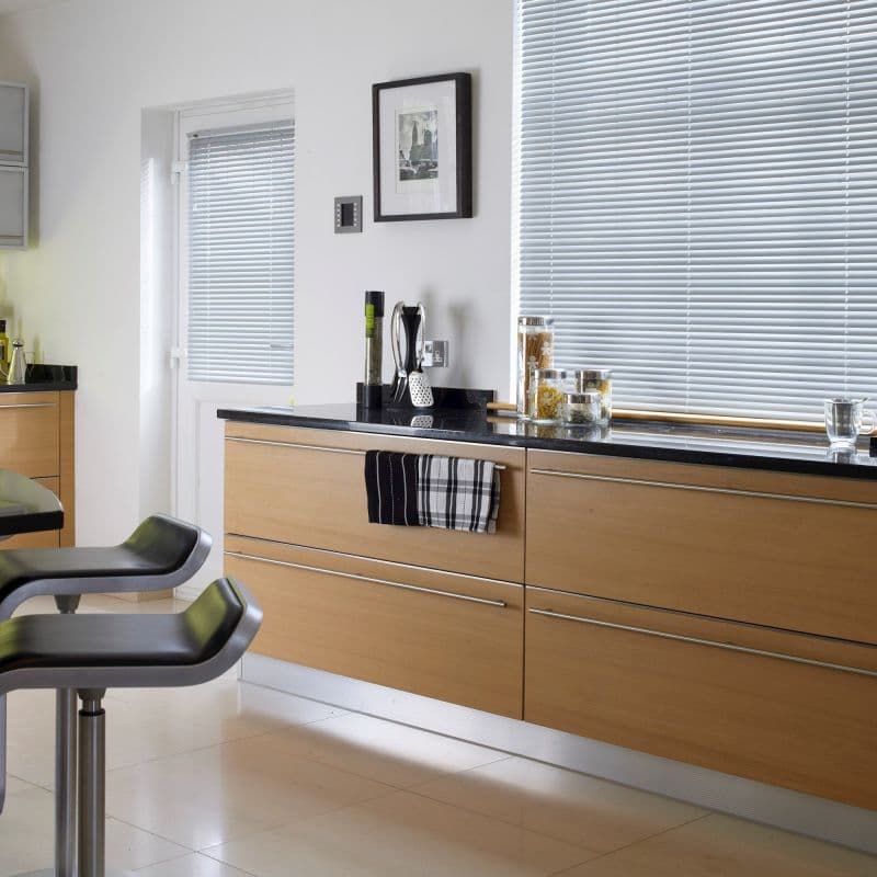 Horizontal Venetian blinds covering a wide window and door, fully closed and filtering daylight; bright modern kitchen with light wood cabinets, black countertop, bar stools and glass jars.