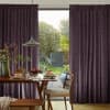 Heavy pleated purple curtains, drawn partially aside to reveal a glass sliding door; curtains frame and filter daylight into a dining area with wooden table, pendant lamp, and garden beyond.