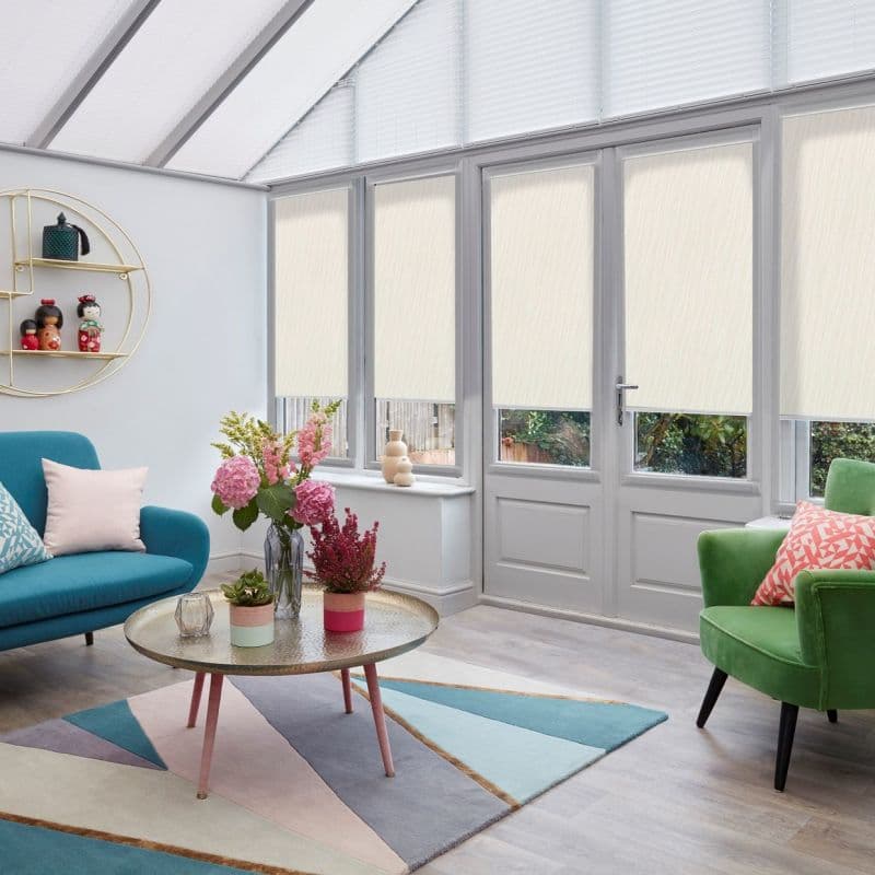 Cream roller blinds cover four tall windows and a door, pleated blinds on triangular roof panes—closed, gently filtering daylight; bright conservatory with teal sofa, green armchair, coffee table and flowers.