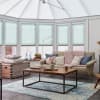 Teal pleated honeycomb blinds covering tall bay/conservatory windows and roof panels, lowered and softly filtering daylight; conservatory living room with beige sofa, pink chunky throw, wooden coffee table and rug.