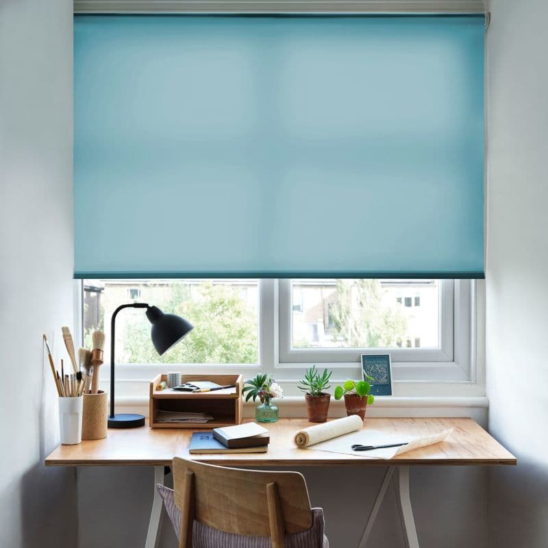 A teal roller blind, lowered over a double window, translucent with a soft gradient, filtering daylight; above a wooden desk in a workspace with lamp, brushes, potted plants and notebooks.