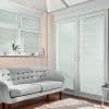 White pleated blinds on French doors and upper windows, fully lowered and producing narrow striped shadows—filtering sunlight into a bright living room with a pale tufted sofa, hardwood floor, and rug.