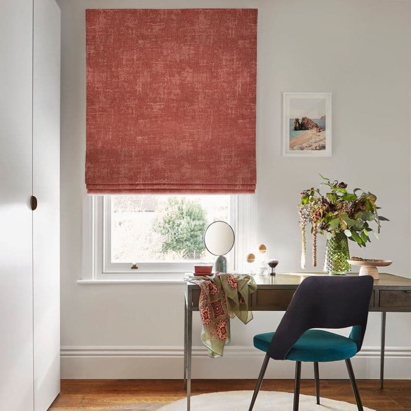 Textured terracotta Roman blind lowered over the window, softly filtering daylight; compact home office/vanity with a desk, teal chair, round mirror, vase of flowers and pale walls.