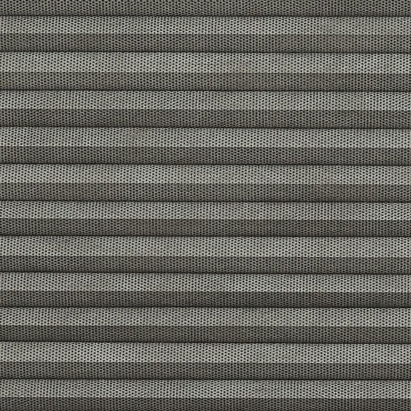 Grey pleated cellular blind, fully closed, narrow horizontal folds with perforated textured fabric; filtering light and covering a window in a softly lit interior, no visible view.