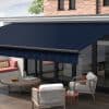 A navy-blue fabric retractable awning with subtle diagonal stripes and matching valance, extended over glass patio doors, shading a sunlit outdoor seating area with table, chairs, brick wall and tree.