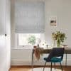 A patterned white Roman blind lowered over a sash window, filtering soft daylight; small home desk beneath with mirror, vase of flowers and teal chair in a neutral, sunlit study.