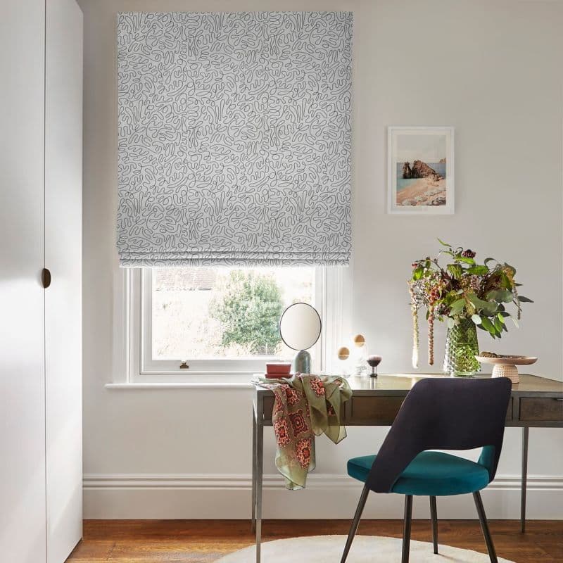 A patterned white Roman blind lowered over a sash window, filtering soft daylight; small home desk beneath with mirror, vase of flowers and teal chair in a neutral, sunlit study.