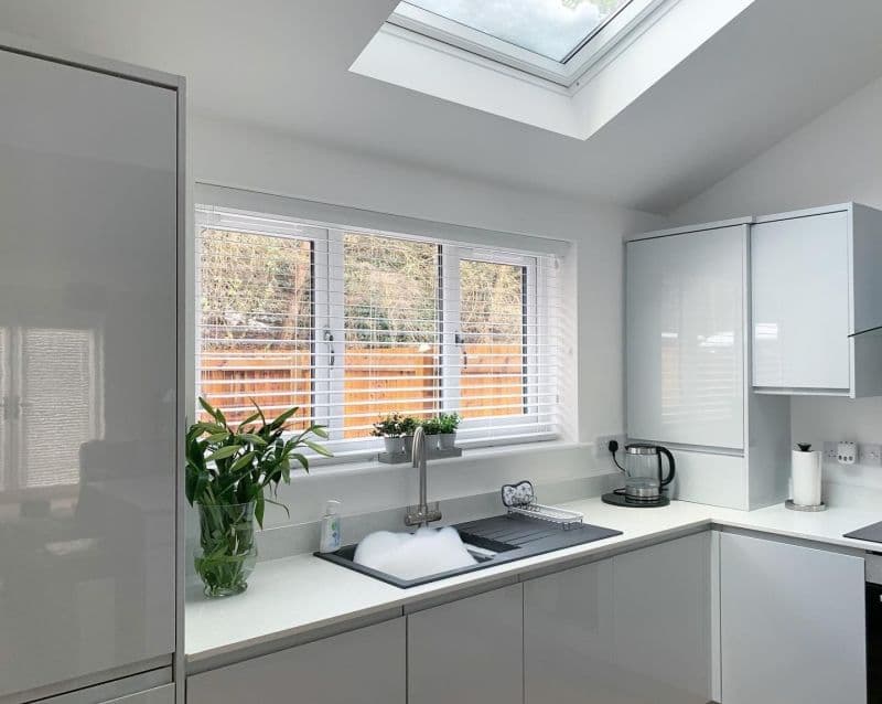 Horizontal white Venetian blinds covering a three-panel window, lowered with slats slightly open, filtering daylight over a modern white kitchen sink filled with soapy water; skylight overhead, fenced yard view.