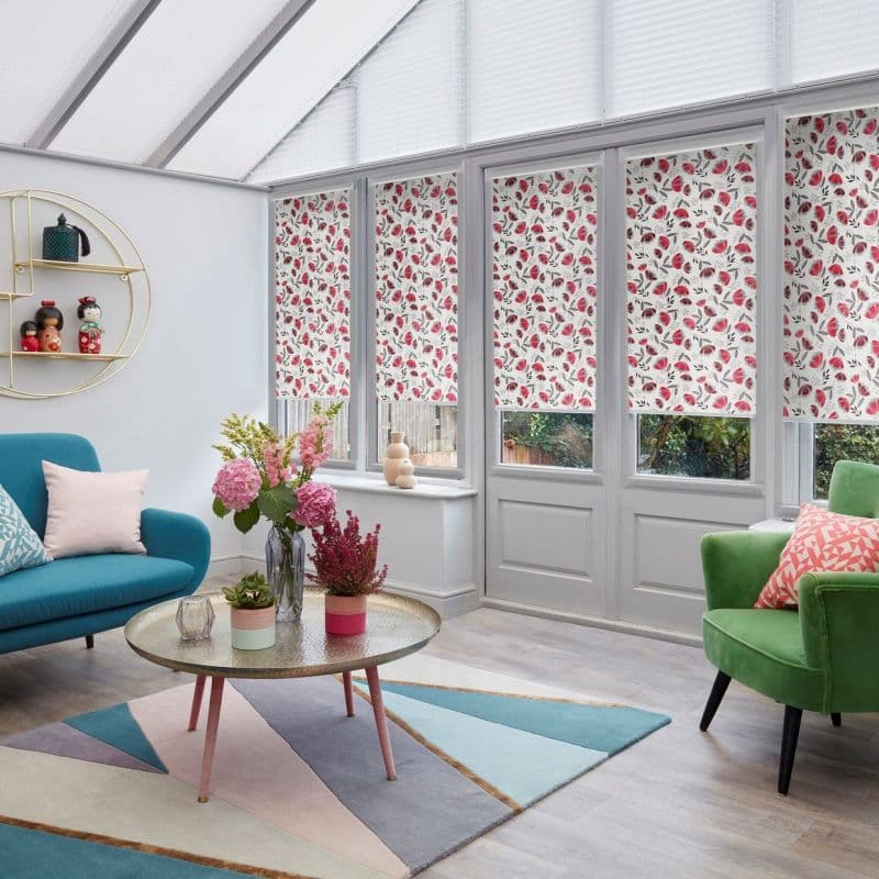 Floral roller blinds, white with red and pink leaves, lowered across tall conservatory windows, mostly closed leaving narrow bottom panes exposed; bright sunroom with colorful seating and a flowered table.