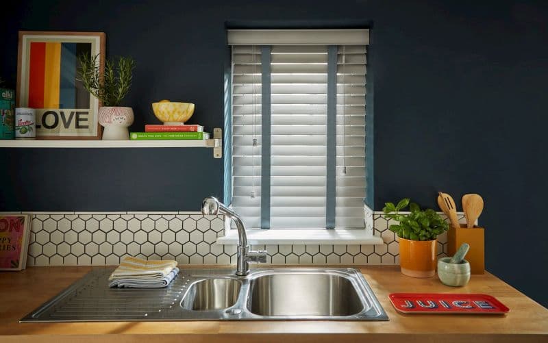 White horizontal Venetian blinds with blue fabric tapes, lowered and partially closed, filtering daylight through slats; kitchen sink with dark blue walls, wooden countertop, hex-tile backsplash, plants and utensils.
Text: LOVE
Text: JUICE
Text: ROSA'S THAI CAFE THE VEGETARIAN COOKBOOK
Text: CRAFT BEER