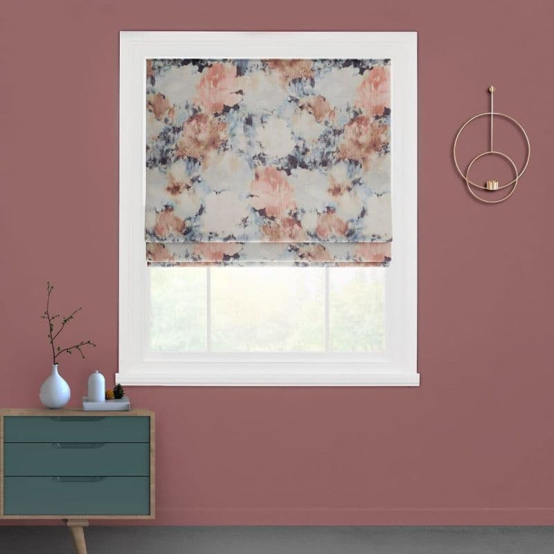 Floral-patterned Roman shade, partially lowered with visible soft folds, diffusing daylight; installed in a white-trimmed window set in a mauve wall, above a teal dresser and gold circular wall sconce.