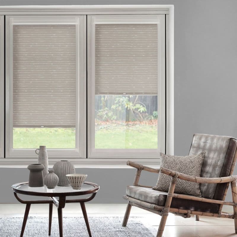 Beige patterned roller blinds (horizontal wavy stripes) covering two windows—one lowered nearly to the sill, the other halfway—partially filtering daylight in a grey living room with rattan armchair and table.