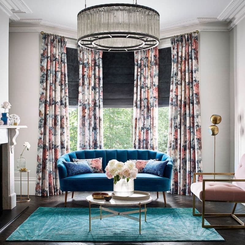 Floral patterned floor-length curtains on a brass rod, pleated and framing bay windows with dark Roman shades partially lowered, filtering daylight; elegant living room with teal sofa, chandelier, leafy view.