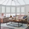White pleated cellular blinds lowered across tall conservatory windows, filtering soft daylight; modern sunroom with beige sofa, chunky pink throw, wooden coffee table, and blue patterned rug.