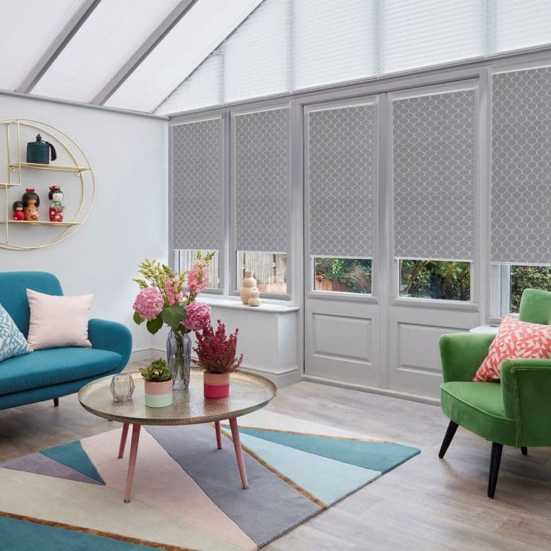 Grey patterned roller blinds, lowered just above the lower panes, partially filtering daylight on French doors and windows in a bright conservatory with mid-century furniture and floral arrangements.