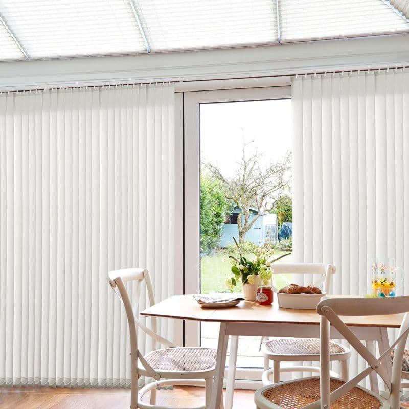 White vertical blinds, floor-to-ceiling, parted at center; filtering daylight and partially open to a glass sliding door; sunlit dining area with wooden table, chairs, and garden view.