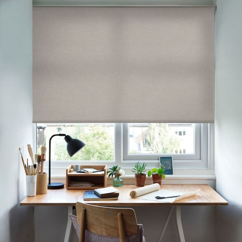 A beige textured roller blind, lowered almost to the sill, partially blocking and diffusing daylight — above a tidy light-wood desk with lamp, brushes, notebooks and small potted plants; blurred trees outside.