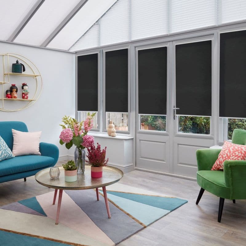 Black roller blinds covering tall conservatory windows and door, mostly lowered to block light; modern sunroom with skylight, colorful armchairs, geometric rug, round coffee table with flowers, exterior garden view.