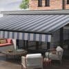 Striped blue-and-white retractable awning extended over a patio, casting shade; patio contains cushioned seating, round wooden coffee table with drinks, glass doors and brick house exterior under daylight.