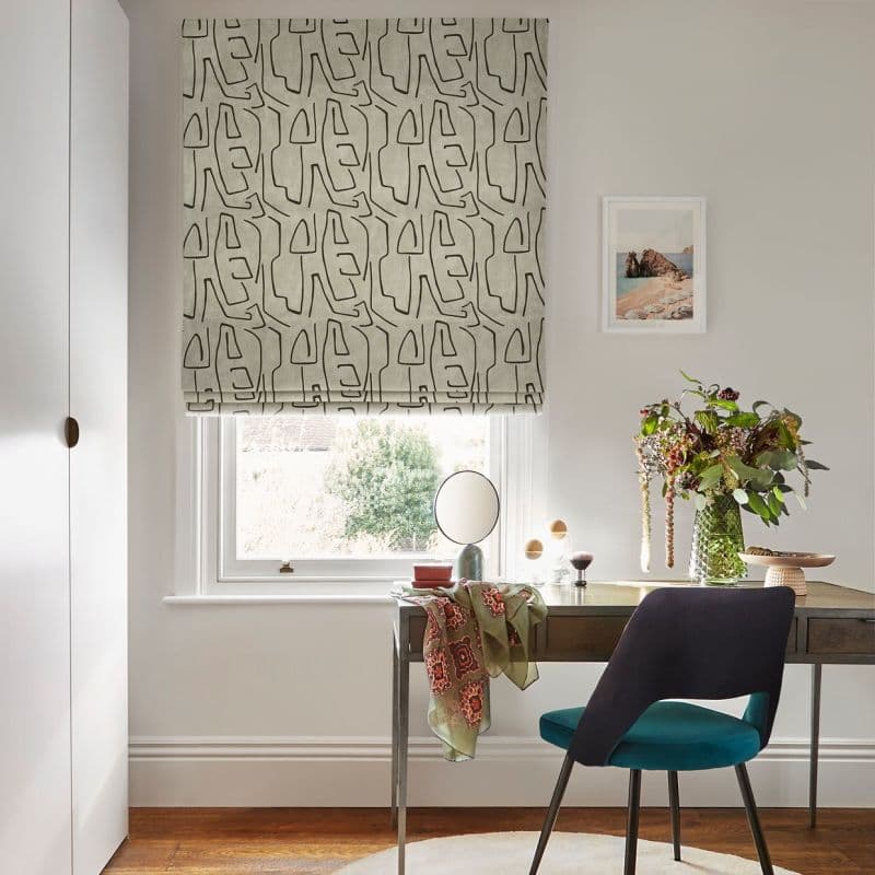 Roman blind—beige fabric with bold black abstract-line pattern—lowered over a window, filtering daylight; small desk and teal chair beneath, vase of flowers, pale-walled home office.