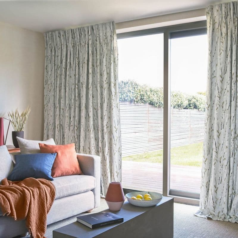 Floor-length pleated curtains with pale botanical print, partly drawn and filtering daylight through sliding glass doors; beige sofa with cushions and coffee table in a sunlit living room.