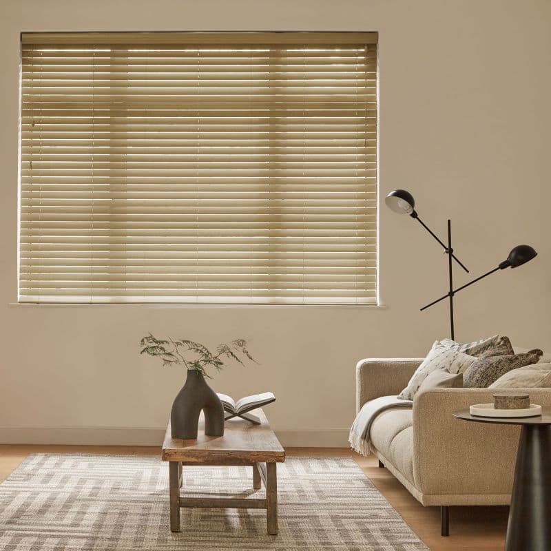 Beige horizontal venetian blinds covering the window, slats mostly closed and filtering soft daylight; above a neutral living room with beige sofa, wooden coffee table and a black floor lamp.