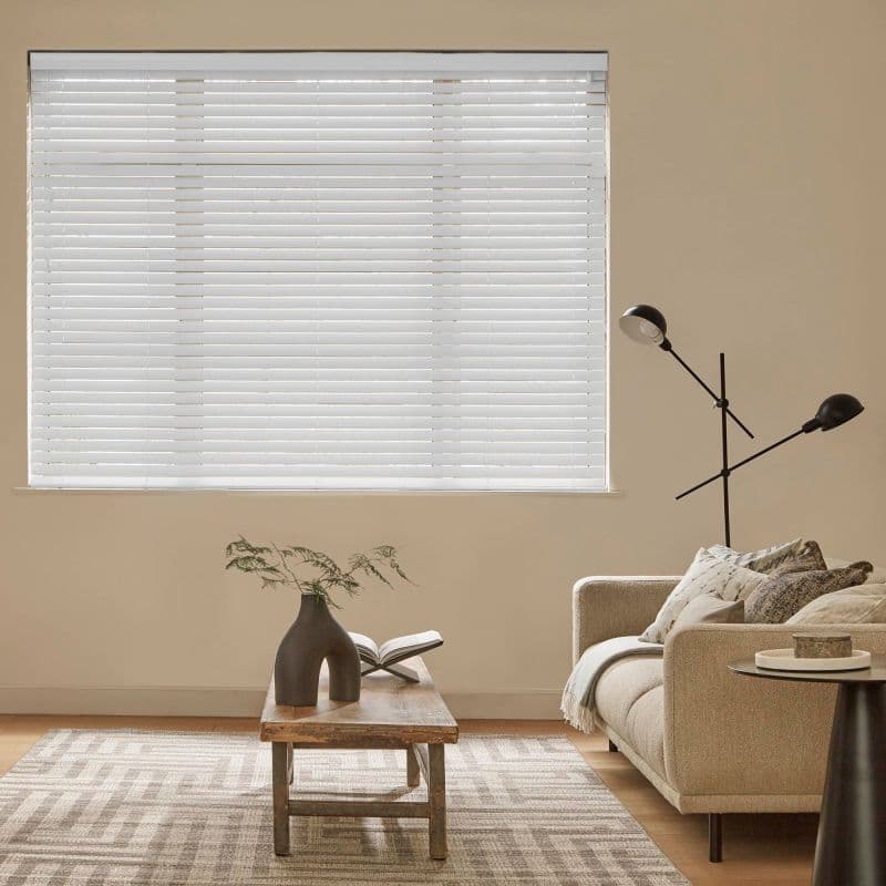 White horizontal blinds covering a rectangular window, closed and filtering soft daylight, in a beige living room with a sofa, wooden coffee table holding a vase and open book.