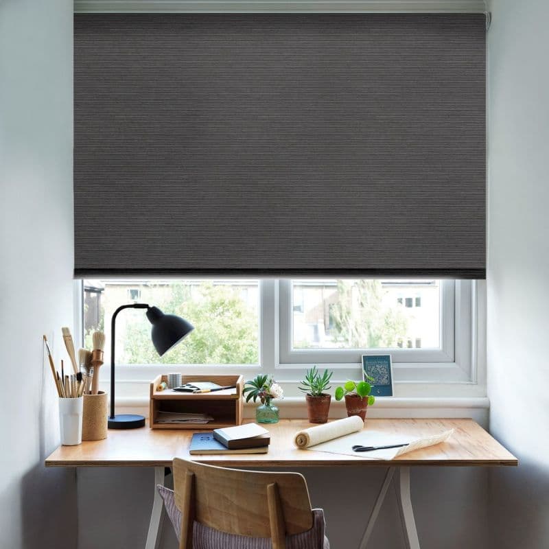 Dark grey fabric roller blind, lowered almost fully with a narrow bottom gap, blocking light; above a wooden desk holding a lamp, art supplies and potted plants in an office.