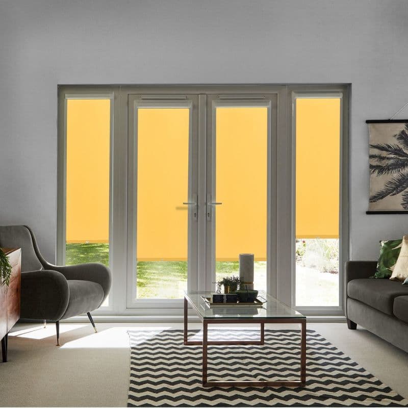 Yellow roller blinds lowered over four-panel patio doors and sidelights, partially blocking light; living room with armchair, sofa, glass coffee table and chevron rug, daylight lawn visible below blinds.