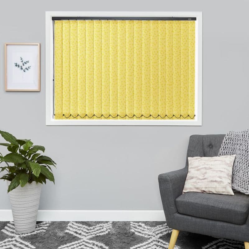 Yellow patterned vertical blinds covering the window — narrow slats with leaf print and scalloped beaded hem — fully closed, diffusing light in a gray living room with plant, armchair, framed print.