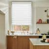 A white zebra roller blind, lowered to cover most of the window with alternating sheer and opaque stripes, gently filters daylight; kitchen with brass sink, wooden cabinets and marble counters.