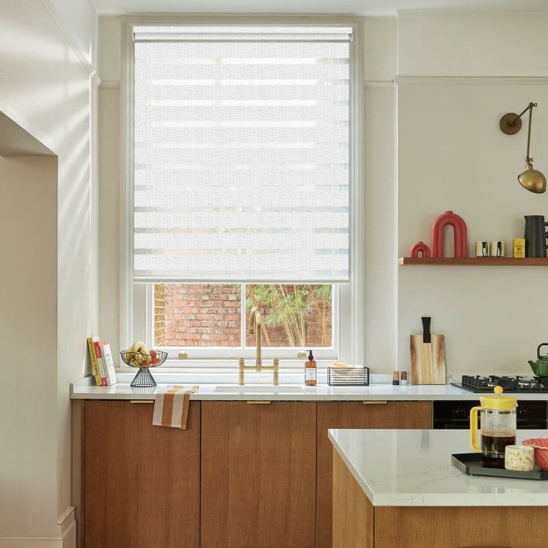 A white zebra roller blind, lowered to cover most of the window with alternating sheer and opaque stripes, gently filters daylight; kitchen with brass sink, wooden cabinets and marble counters.