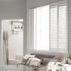White louvered plantation shutters (three-panel): left panel folded open, right panels closed, partially filtering bright daylight over a bench with neutral cushions and throws in a light, coastal-style sitting area.