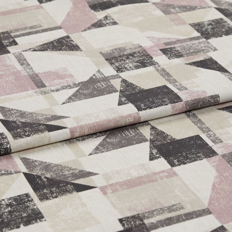 Folded upholstery fabric with distressed geometric triangles and rectangles in muted pink, beige and charcoal — lying layered on a flat surface, close-up under soft studio lighting.