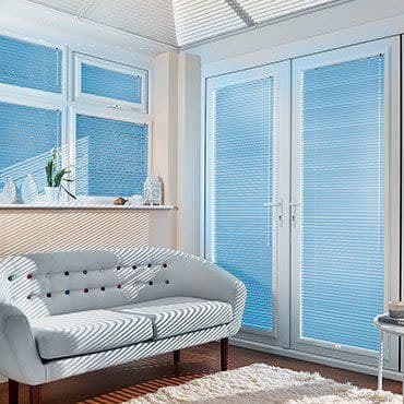 White horizontal blinds on bay windows and French doors, closed but filtering sunlight; striped light falls across tufted white loveseat in bright living room with wooden floor and rug.