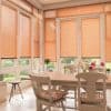 Peach-colored horizontal Venetian blinds cover tall glass doors and windows, mostly lowered and filtering warm daylight; dining nook with a wooden table, chairs with floral cushions, vases, and garden view.