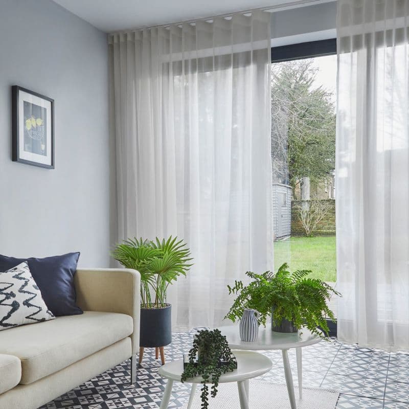 Sheer, floor-to-ceiling white curtains hung on a ceiling track, partially drawn and softly filtering daylight across a tiled living room with sofa, houseplants, and a grassy garden visible outside.