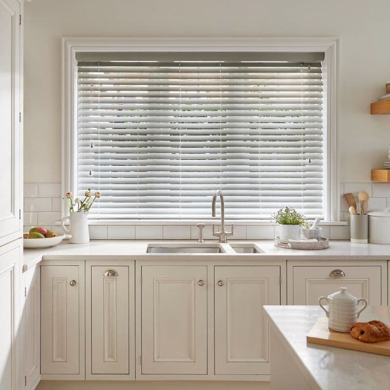 White horizontal Venetian blinds cover a large kitchen window; slats mostly closed, angling to filter soft daylight, above a white sink and marble countertop in a bright, neutral kitchen.