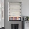 A beige zebra roller blind covers a recessed window, alternating opaque and sheer horizontal bands partially filtering light; set in a gray kitchen with espresso machine, radiator and framed art.