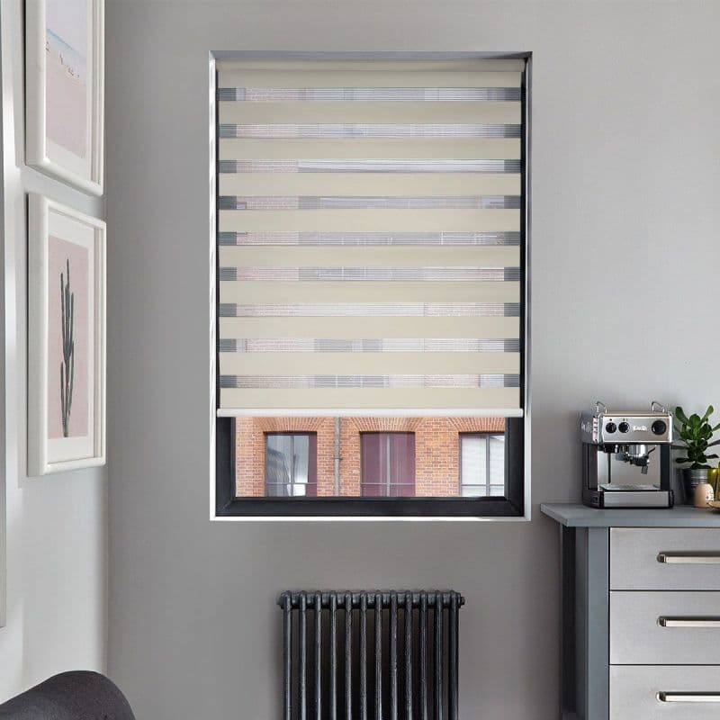 A beige zebra roller blind covers a recessed window, alternating opaque and sheer horizontal bands partially filtering light; set in a gray kitchen with espresso machine, radiator and framed art.