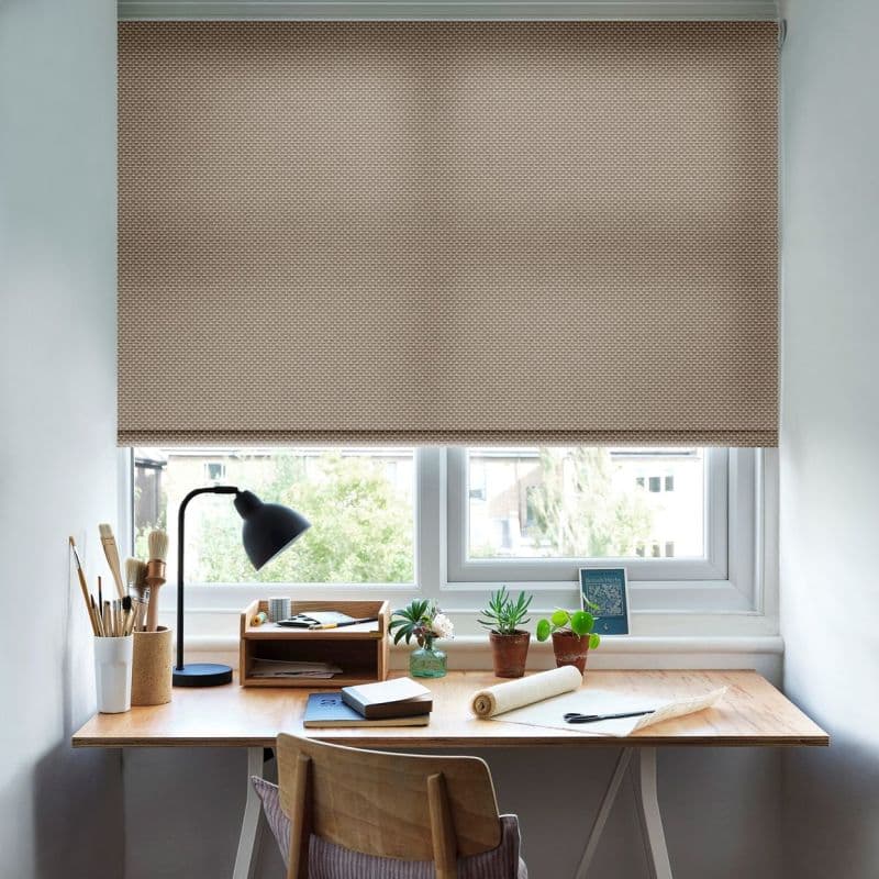 A beige textured roller blind, mostly lowered across the window, filtering daylight; above a wooden desk with black lamp, potted succulents, paintbrushes, notebooks, and a chair in a bright room.