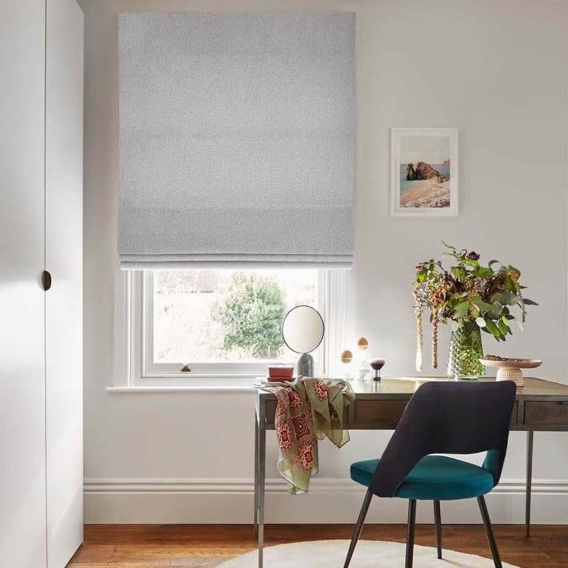 Grey patterned Roman blind lowered over window, filtering soft daylight; a wooden desk with mirror, vase of flowers and draped scarf; bright home office with neutral walls and teal chair.